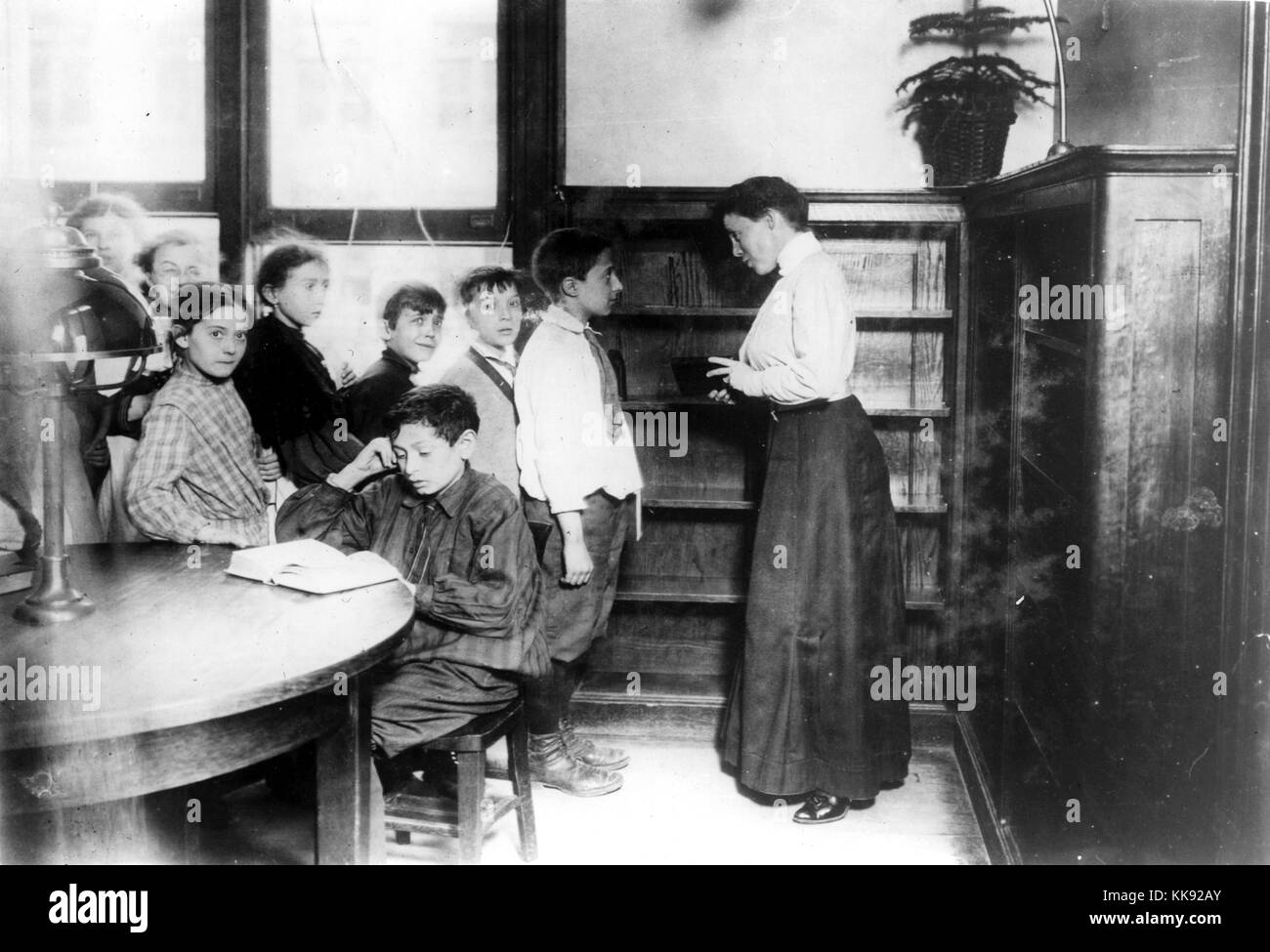 A group of students are gathered to listen to the librarian at the Seward Park Library, most of the students are distracted by the photographer, a student sits at the table reading, the shelves behind the librarian are empty, New York, New York, 1910. From the New York Public Library. Stock Photo