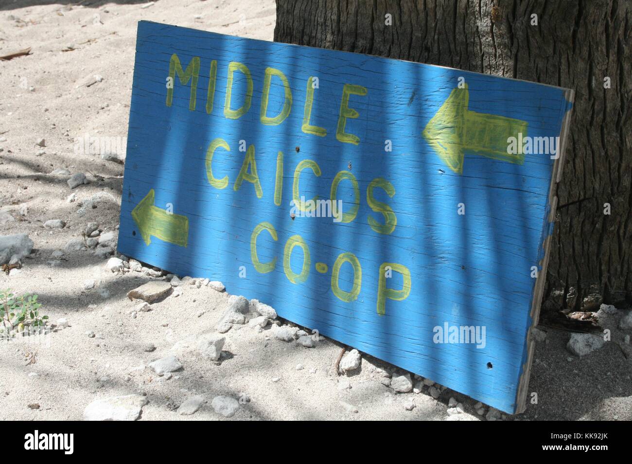 A photograph of a painted wooden sign that reads 'Middle Caicos Co-Op', the blue sign also has arrows that point in the direction of the business, it rests against a tree in the sand, the store represents local artists that produce souvenirs for tourists, Turks and Caicos Islands, 2013. Stock Photo