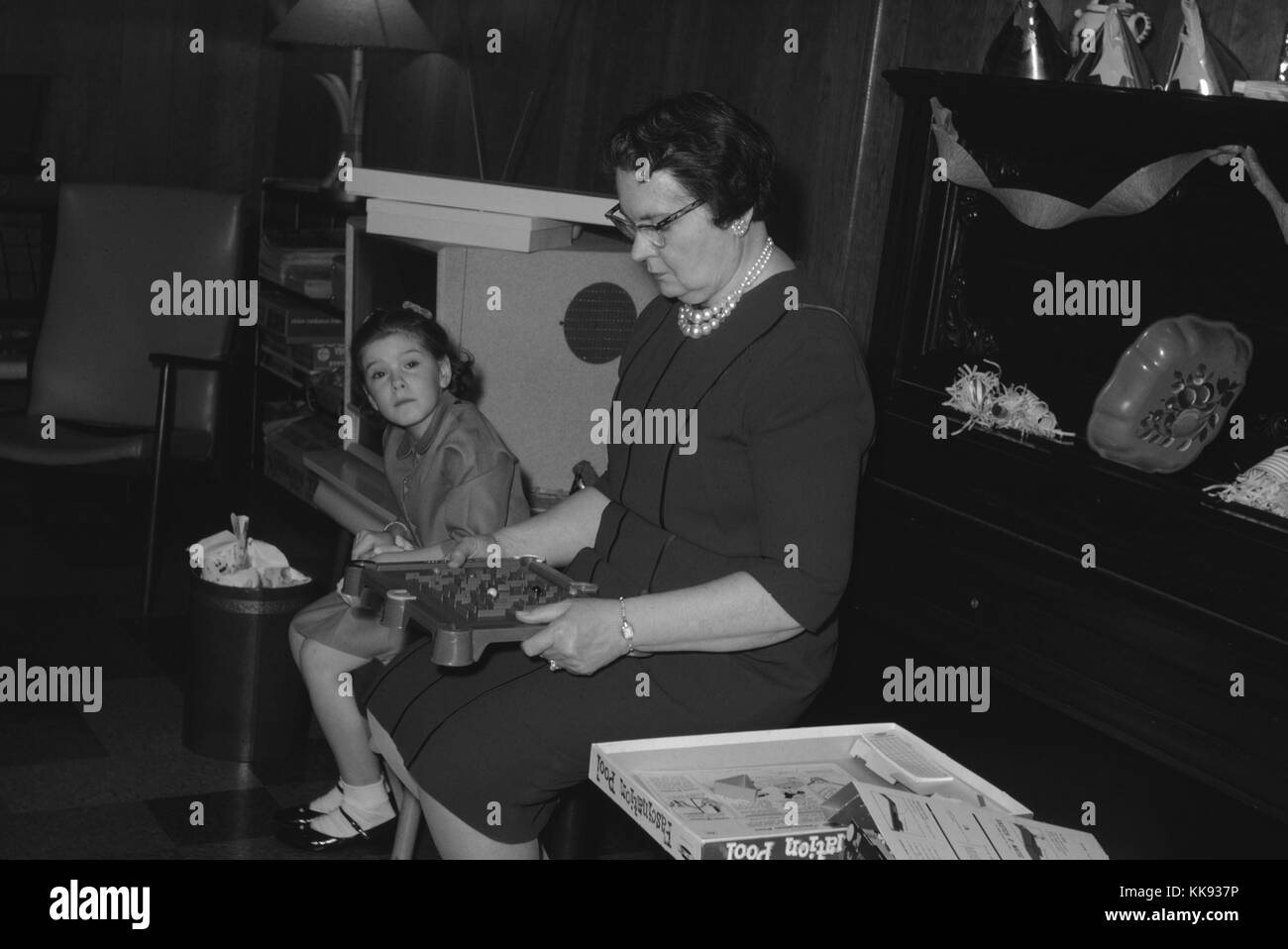 Grandmother in a blue dress sitting upright and playing with a marble board game during a party, while her granddaughter looks on, 1963. Stock Photo