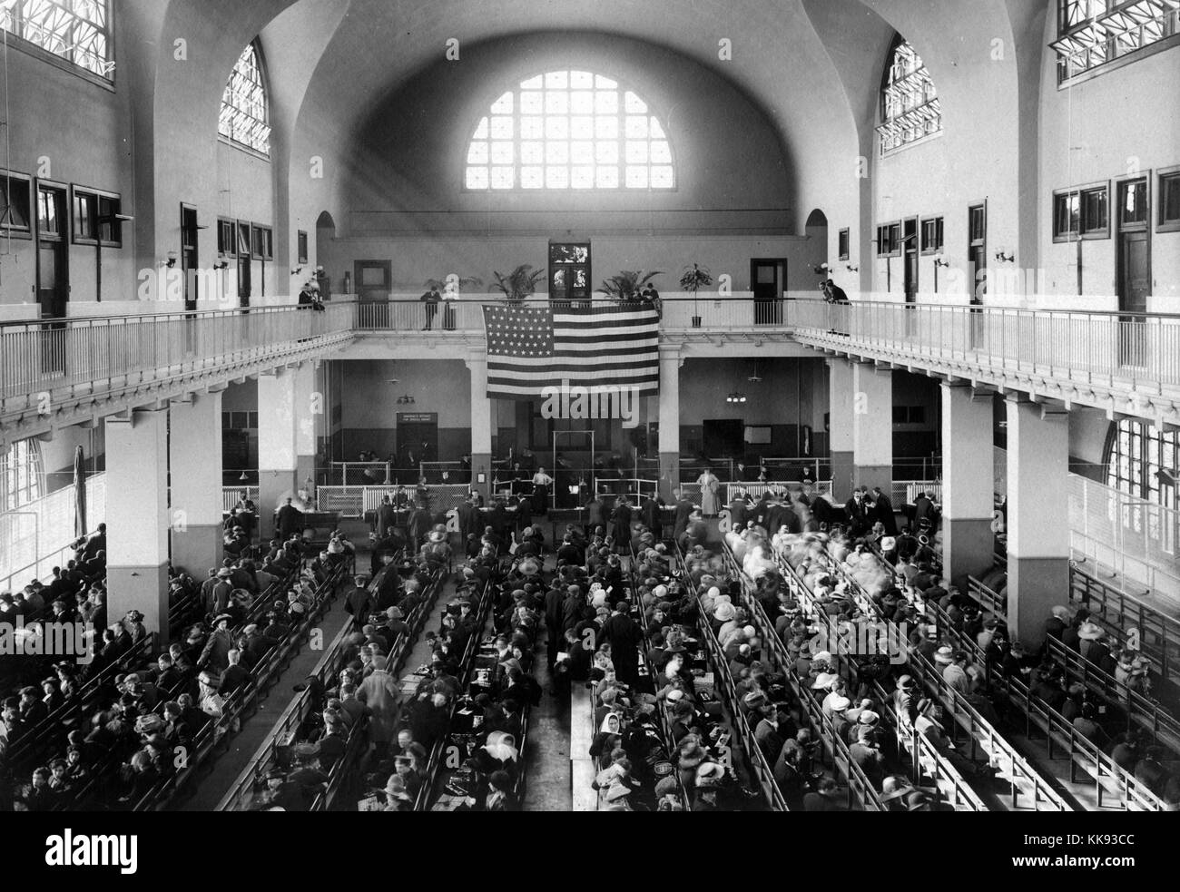 A photograph of the main hall of the United Station immigration inspection station on Ellis Island, immigrants line the benches of the main hall while a few people can be seen looking down at the crowd from the second floor, the American Flag hanging over the main hall displays 46 stars, Ellis Island was the busiest point of immigration in the United States from 1892 until it closed in 1954 after processing over 12 million immigrants, New York, 1907. From the New York Public Library. Stock Photo