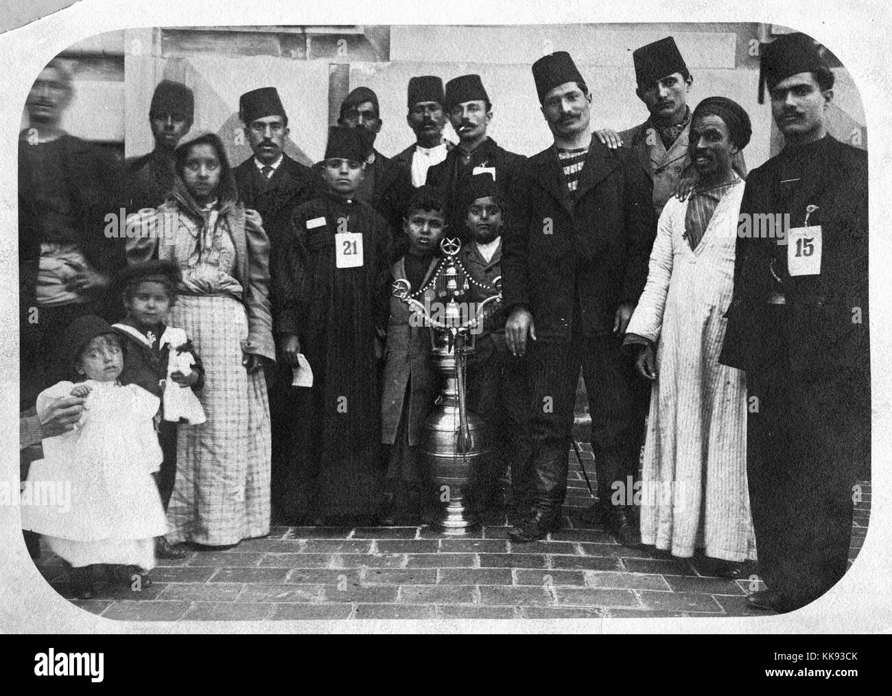 A photograph of a group of immigrants on Ellis Island, most of the men and some of the boys are wearing fezzes, there are several young children in photo, at the center of the group is a large metallic vessel that contains three star and crescent symbols which are used by the Muslim religion and by Ottoman Turks, New York, 1907. From the New York Public Library. Stock Photo