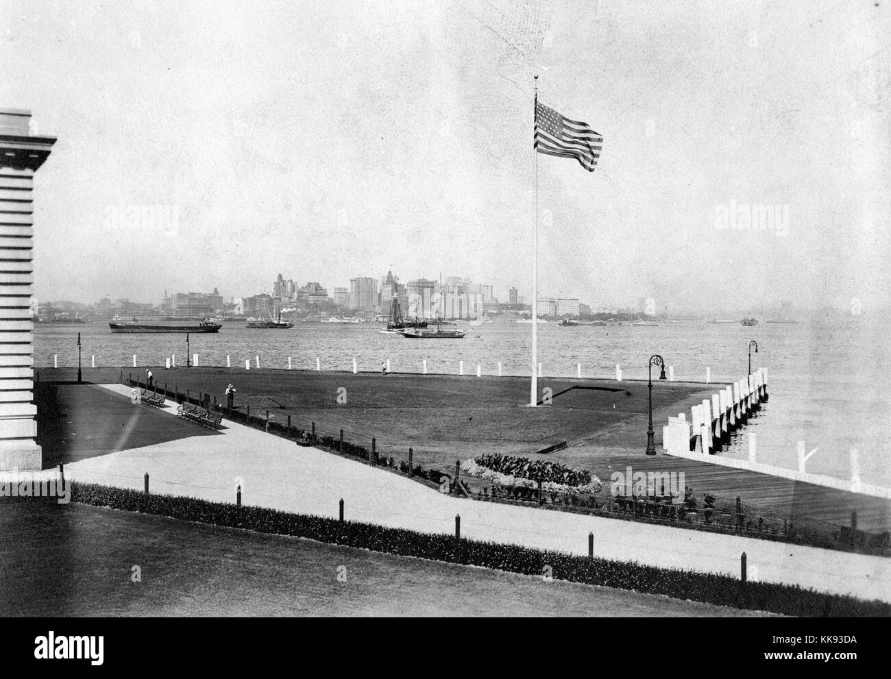 A photograph showing a portion of the lawn that surrounds the immigration inspection station on Ellis Island, the French Renaissance Revival building is constructed with red bricks and limestone trim, the building was first opened on December 17, 1900, it was closed in 1954 after processing over 12 million immigrants entering the United States, a flag is being flown outside of the building, a wide walkway is separated from the grass by low fences and shrubs, New York City can be seen in the background across the Upper New York Bay, 1907. From the New York Public Library. Stock Photo