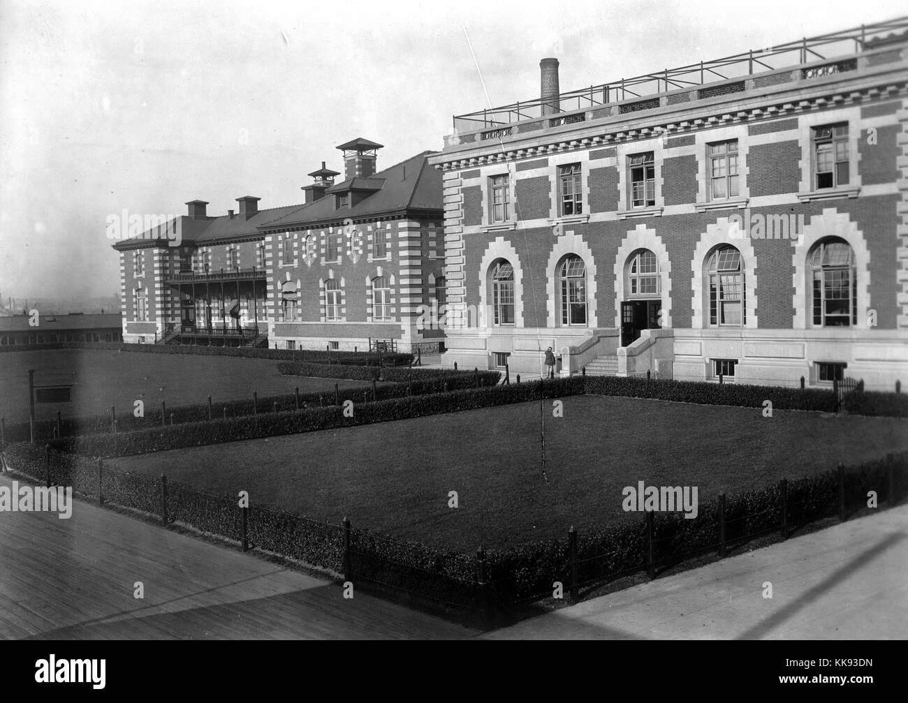 An exterior photograph of a portion of the Ellis Island immigration inspection station, the French Renaissance Revival building is constructed with red bricks and limestone trim, the building was first opened on December 17, 1900, it was closed in 1954 after processing over 12 million immigrants entering the United States, plots of grass are protected by short fences and shrubs, wide walkways surround the grass and building, New York, 1907. From the New York Public Library. Stock Photo