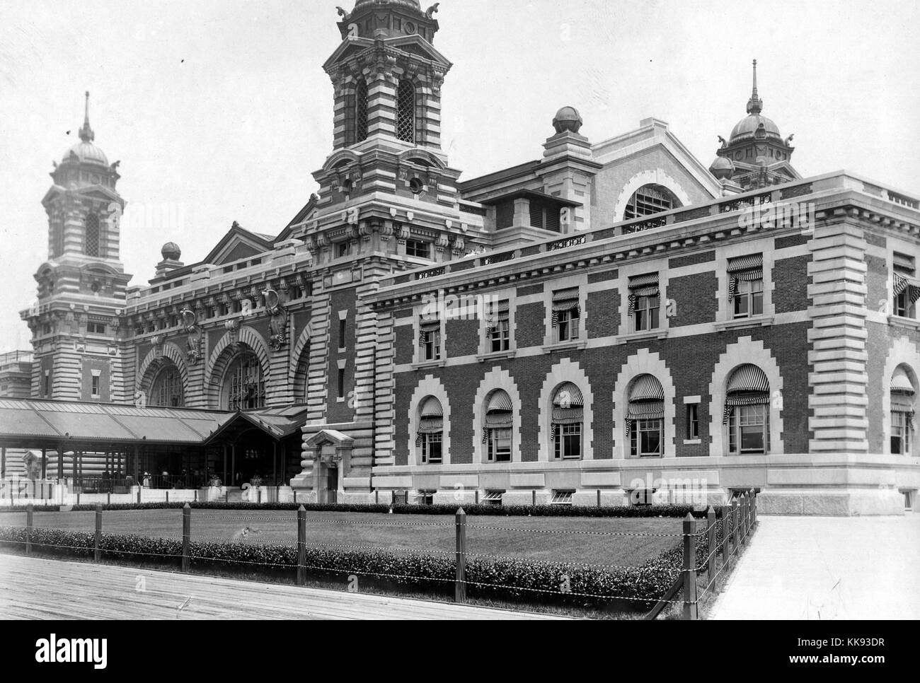 An exterior photograph of a portion of the Ellis Island immigration inspection station, the French Renaissance Revival building is constructed with red bricks and limestone trim, the building was first opened on December 17, 1900, it was closed in 1954 after processing over 12 million immigrants entering the United States, towers can be seen at the corners of the central portion of the structure with a covered walkway leading to main entrance, a large plot of grass is protected by a short fence and shrubs, wide walkways surround the grass and building, New York, 1907. From the New York Public  Stock Photo