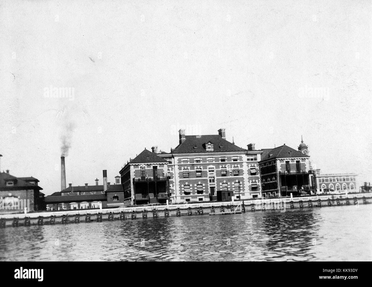 A photograph of the exterior of the Ellis Island Hospital Building as seen from across the water, the hospital was opened in 1902 and operated until 1930, it was the first public hospital in the United States, it was used as a detention center for immigrants who were deemed too ill to enter the country upon their arrival, if the person could not be cured they were sent back to their home country, other buildings including portions of the immigration inspection station and power plant can be seen in the background, New York, 1907. From the New York Public Library. Stock Photo