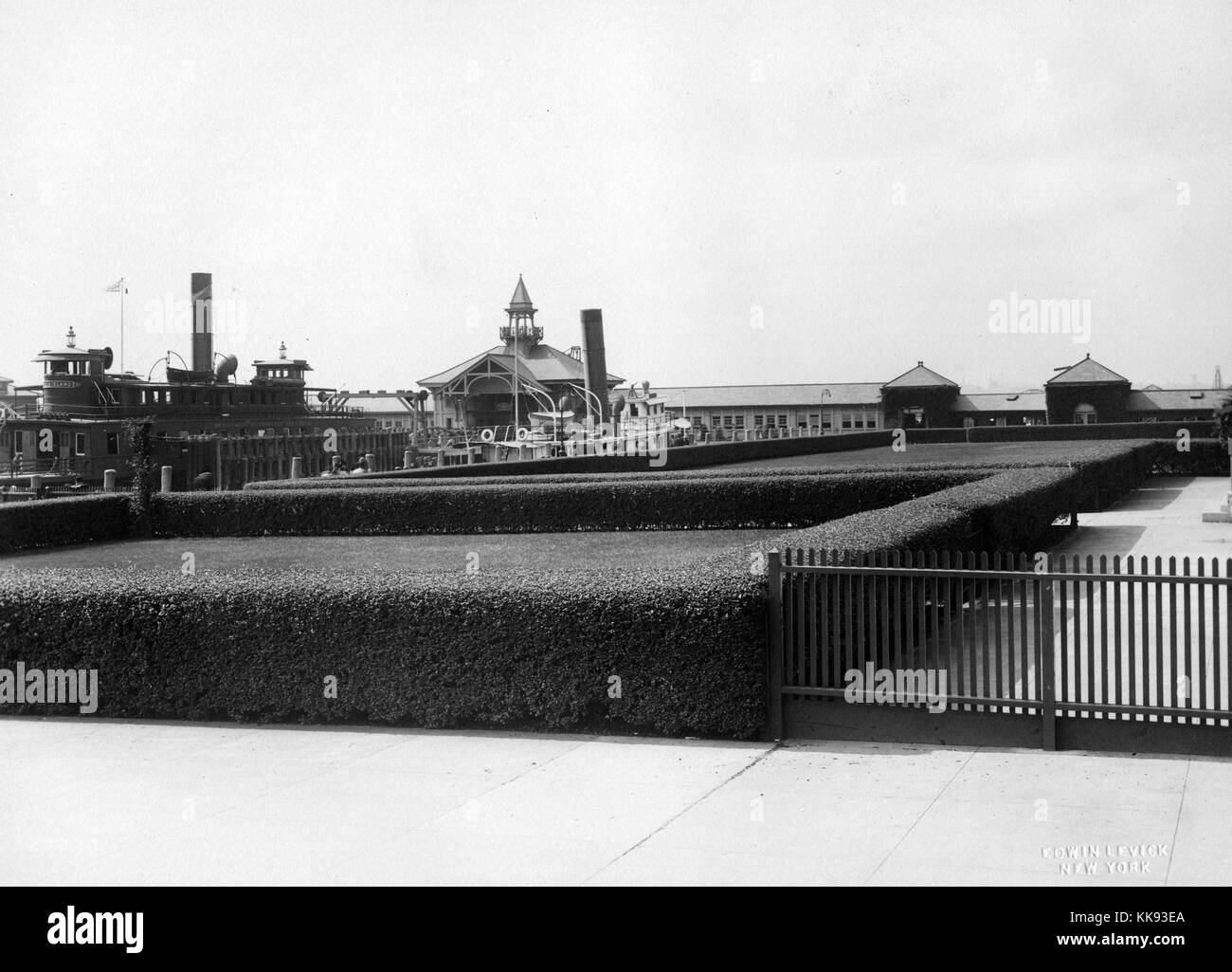 A photograph of the pier on Ellis Island, the ferry docked on the left side of the image was operated by the Department of Commerce and Labor, they oversaw the United States Immigration Services between 1903 and 1913, buildings that make up a small portion of the structures on Ellis Island can be seen in the background, grassy lawns walled off by scrubs are surrounded by large walkways, Ellis Island opened in 1892 and it was closed in 1954 after processing over 12 million immigrants, New York, 1907. From the New York Public Library. Stock Photo