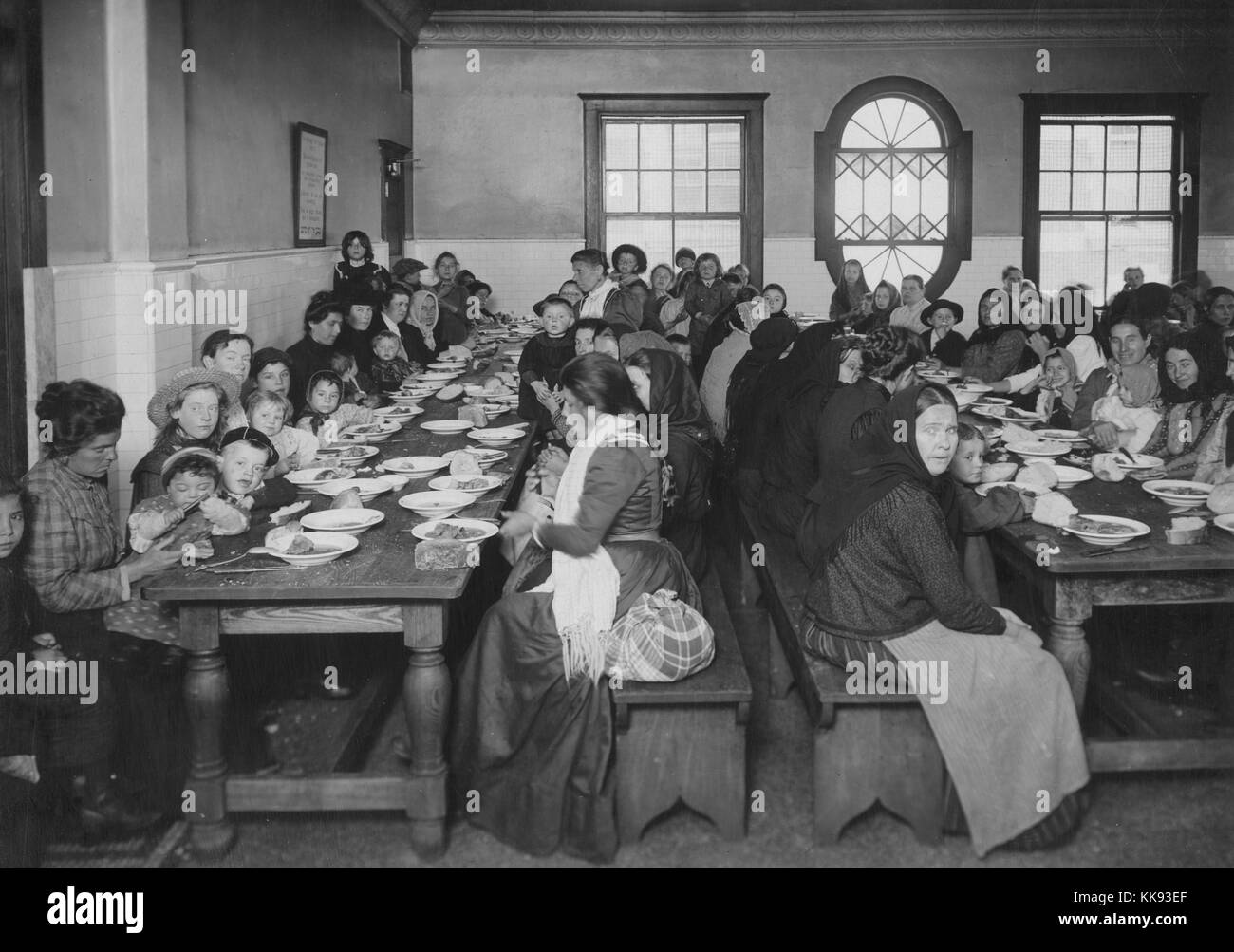 A photograph of a large group of women and children eating a meal on Ellis Island, the long wooden tables are very close together to accommodate as many people as possible in this dining hall, free meals were provided to immigrants and packaged goods were also available for sale to eat between meal times and for immigrants to take away when they left Ellis Island, New York, 1907. From the New York Public Library. Stock Photo