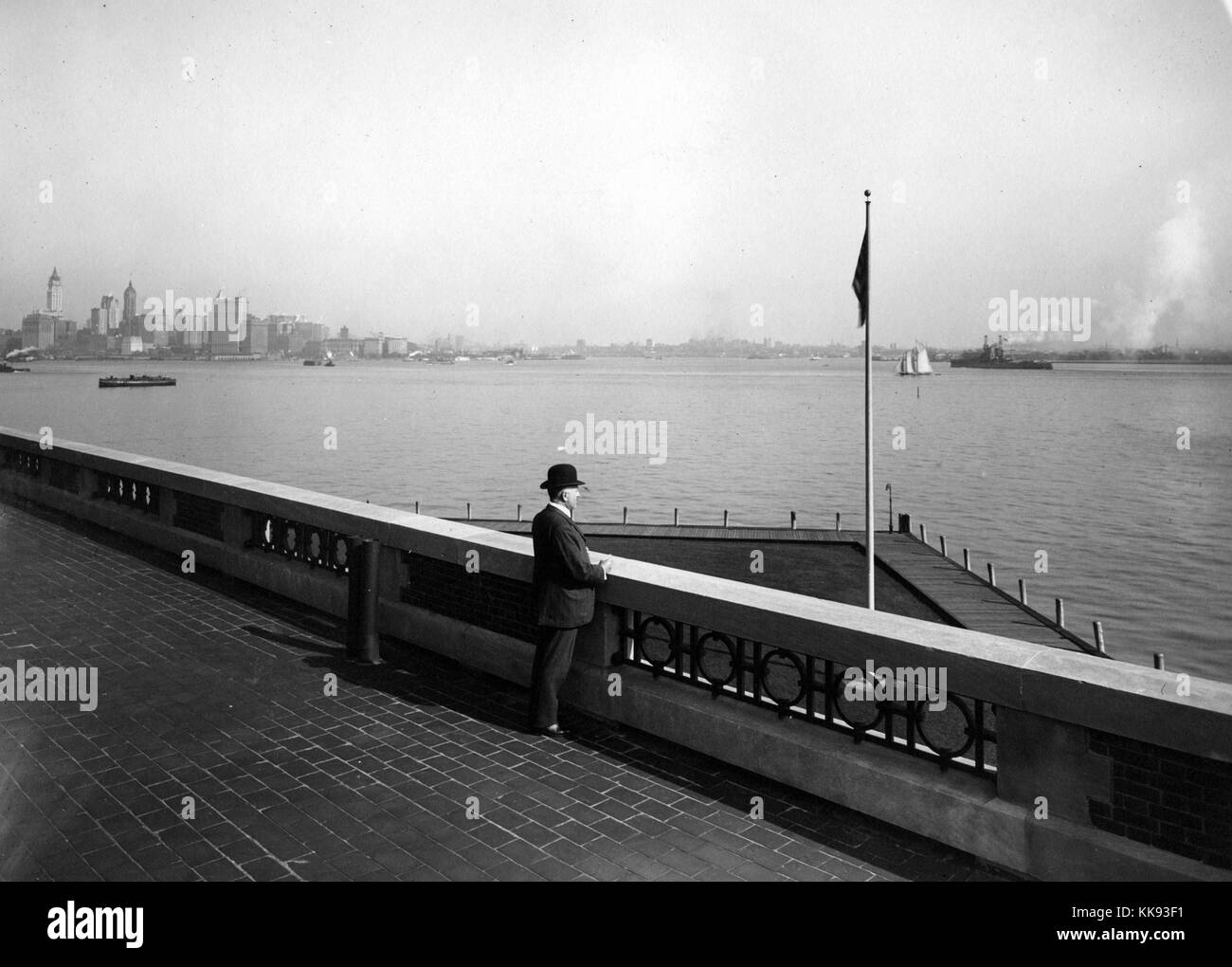 Black and white photograph with a gentleman wearing a suit and derby hat observing the harbor from the observation roof of the Immigration Station, the New York City skyline, showing the nearly completed Woolworth Building Tower, can be seen to the left, by Edwin Levick, Ellis Island, New York, 1907. From the New York Public Library. Stock Photo
