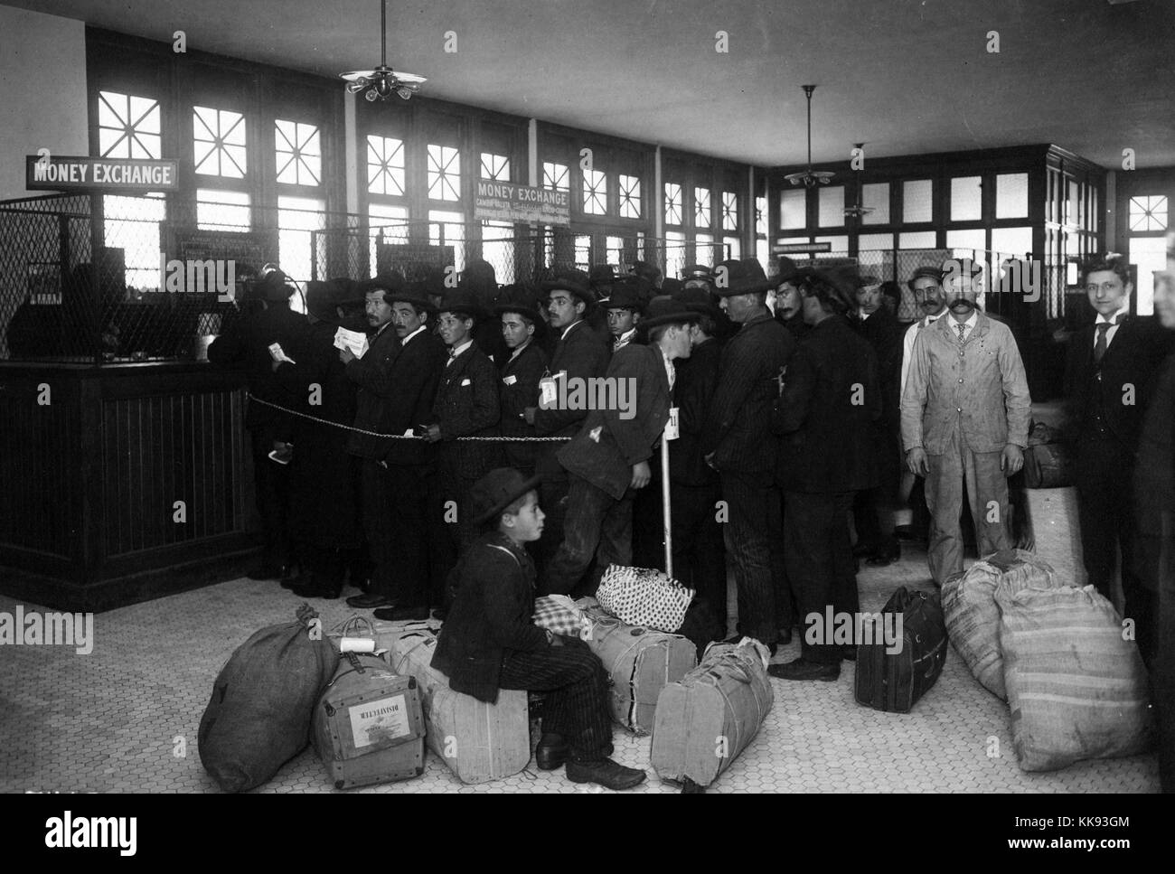 Black and white photograph of a large group of immigrants with baggage lined up at tellers windows marked 'Mone Exchange', by Edwin Levick, Ellis Island, New York, 1907. From the New York Public Library. Stock Photo