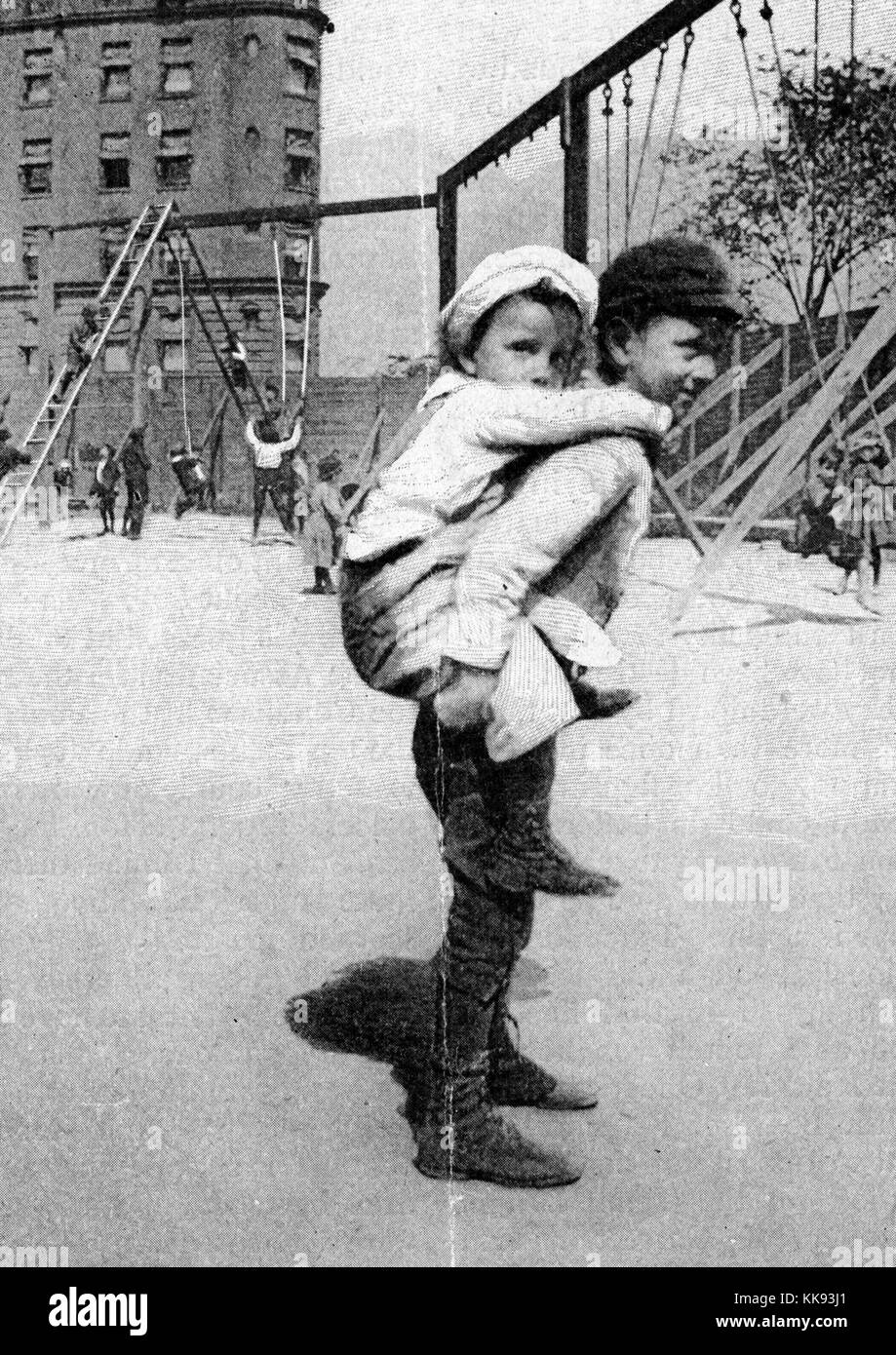 Black and white photograph of a boy carrying his younger brother on his back, at a playground, captioned 'In The New Playground', by Jacob Riis, a Danish-American social reformer and social documentary photographer known for using his photographic and journalistic talents to help the impoverished in New York City, New York City, New York, 1901. From the New York Public Library. Stock Photo