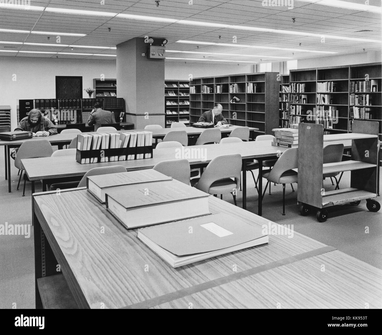 Black and white photograph of a library reading area, with long tables, shelves of reference books, and readers in background, New York City, New York, 1970. From the New York Public Library. Stock Photo