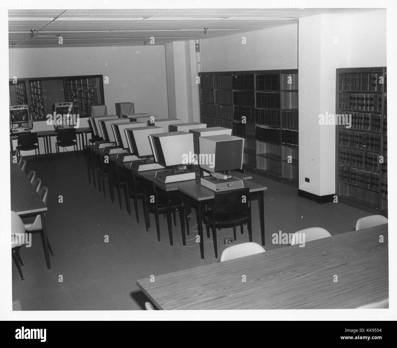 Black and white photograph of a room with a group of microfilm viewers in the center, long tables with chairs, and shelves with books along the walls, New York City, New York, 1970. From the New York Public Library. Stock Photo