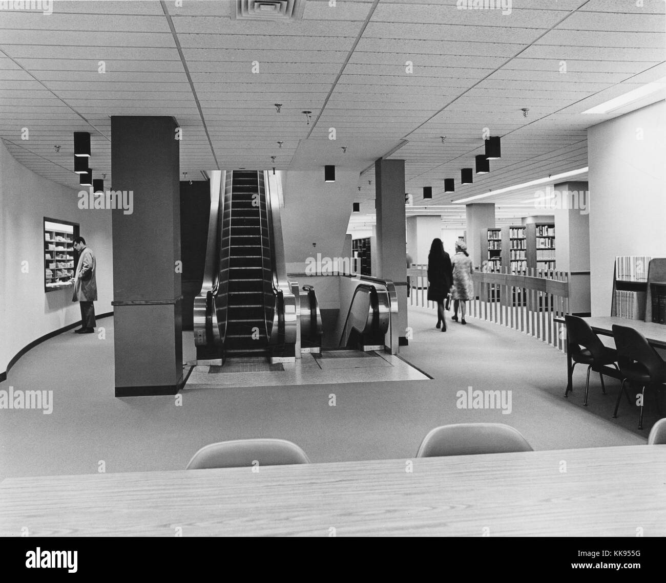 Black and white photograph of an escalator at a library, rows of shelves with books, and people walking can be seen in the background, New York City, New York, 1970. From the New York Public Library. Stock Photo
