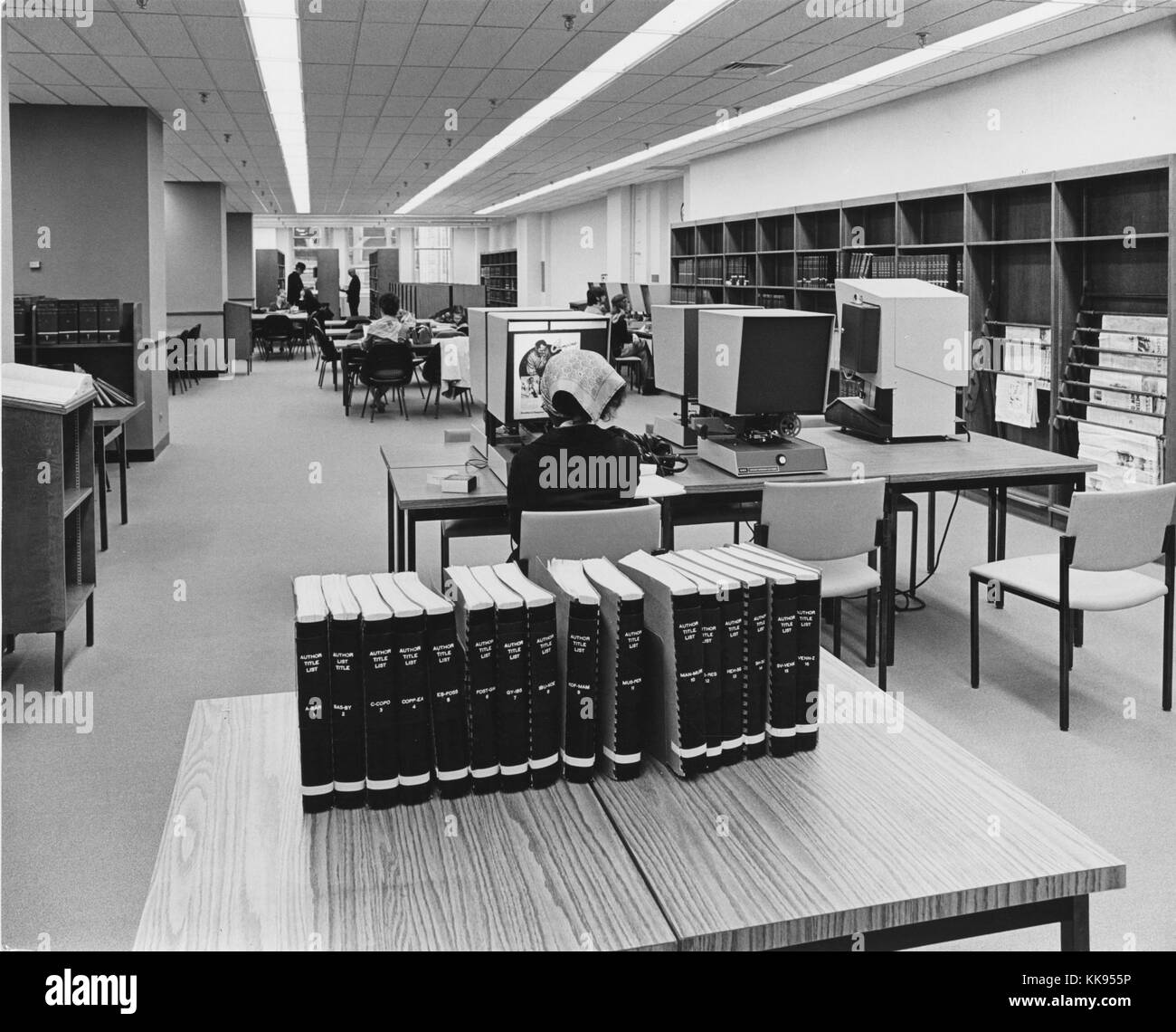 Black and white photograph of a woman sitting at a microfilm viewer, a table with books in the foreground, people sitting at other microfilm viewers in the background, New York City, New York, 1918. From the New York Public Library. Stock Photo