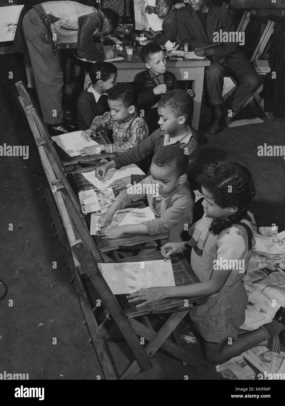 A photograph of children engaging in art training, smaller children are kneeling in front of wooden folding chairs and are drawing on pieces of paper, older children are sharing a desk in the background to work on their art, they are all participating in art training that was sponsored by the Good Shepherd Community Center in Chicago, Illinois, 1877. From the New York Public Library. Stock Photo