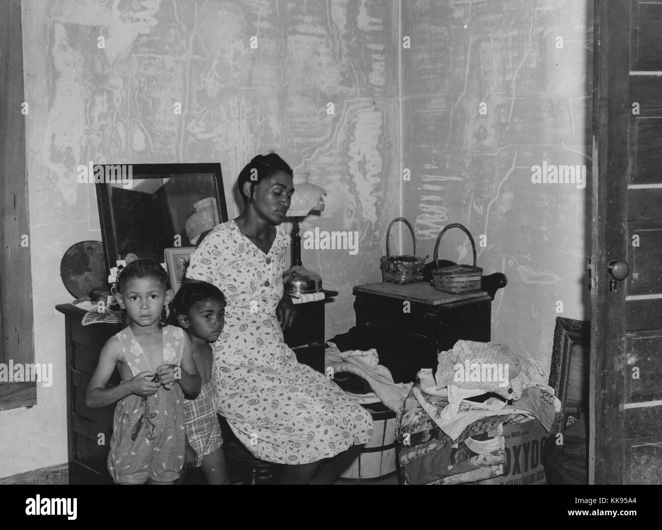 A photograph of a woman and her two children, they are sitting in a room where their belongings can be seen stacked up in boxes, they were among a group of citizens forced to move due to the United States Army taking over their properties to use for training grounds, they were between moves because the Army was also going to take over the area to which they had moved, Caroline County, Virginia, June, 1941. From the New York Public Library. Stock Photo