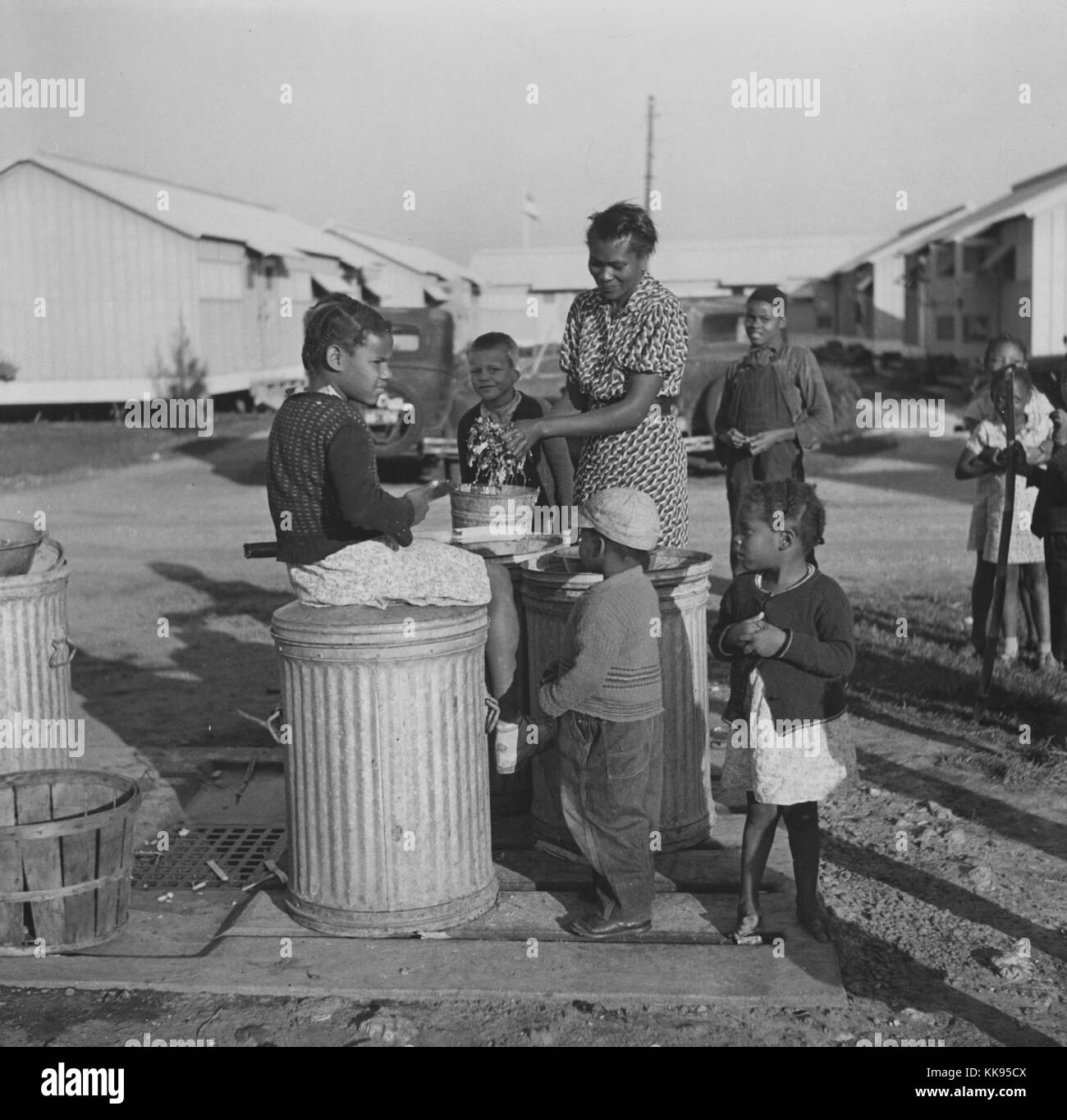 Black and white photograph of an African-American woman, with children, washing greens by pump near metal shelters at Okeechobee Migratory Labor Camp, Belle Glade, Florida, February, 1941. From the New York Public Library. Stock Photo