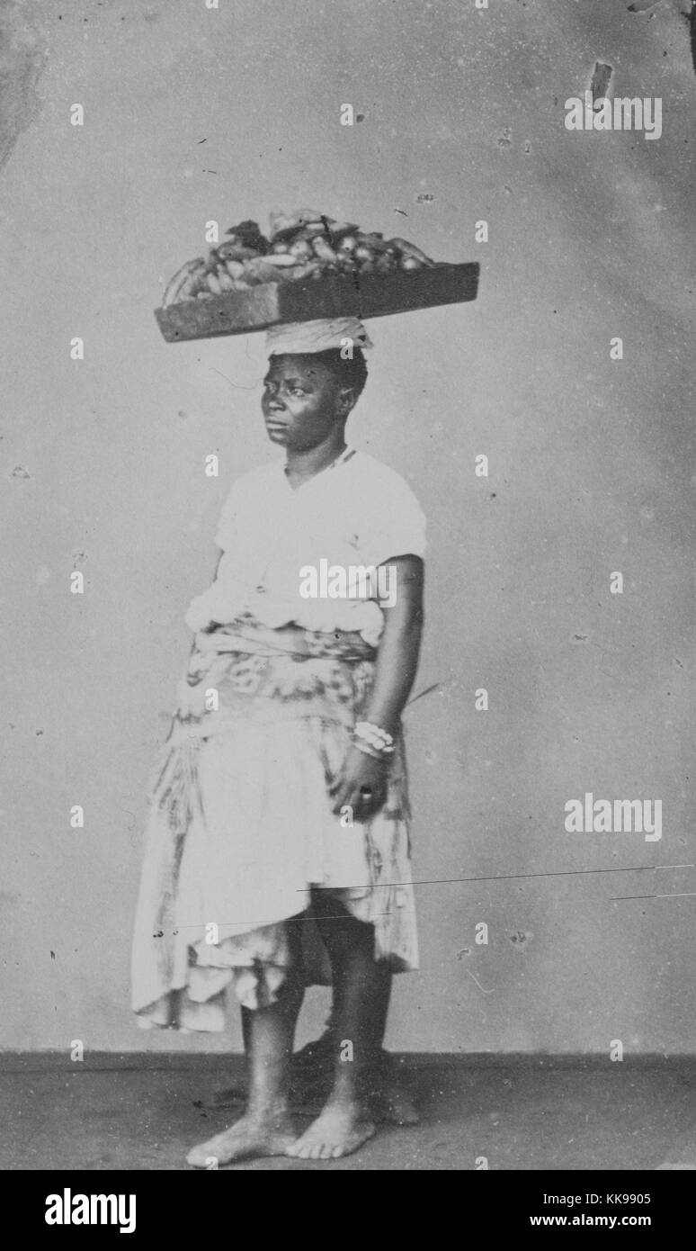 A full length photographic portrait of a woman with a wooden tray on her head, the tray is full of bananas that the woman is taking to a market, she wears a small cloth hat on her head to stabilize the tray, she is wearing a knee length light colored dress, Brazil, 1900. From the New York Public Library. Stock Photo