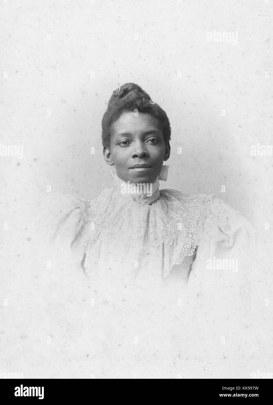 A photographic portrait of a woman wearing a light colored dress, the dress has lace work extending from the neck line to her chest, she has her hair tied up in a bun, 1900. From the New York Public Library. Stock Photo