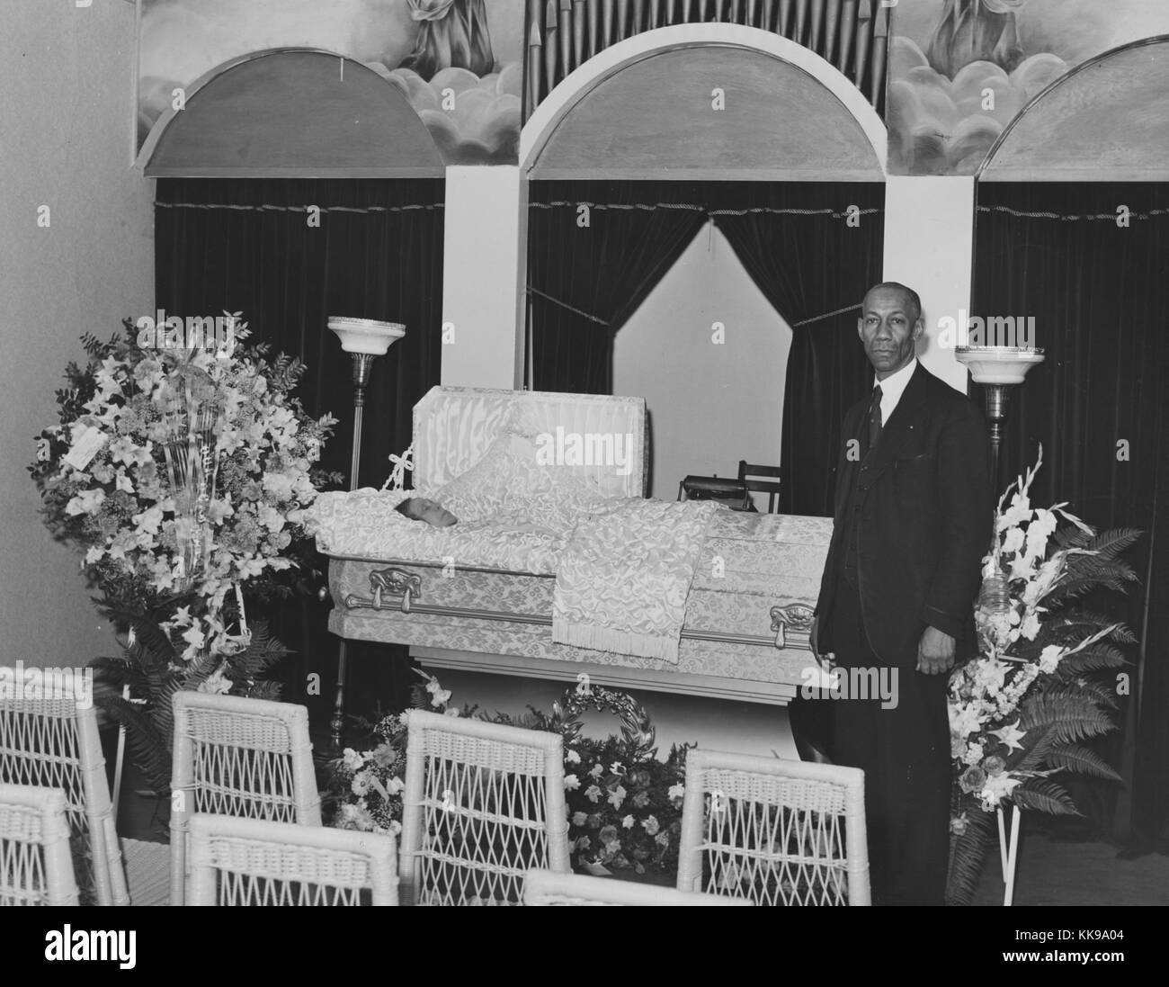 A photograph of an undertaker standing at the foot of a casket. He is wearing a black suit and has a solemn expression on his face. Only the face of the person in the casket is visible. The room has been decorated with bouquets of flowers to prepare it for the funeral service that is about to be held. Religious imagery is painted on the wall surrounding the pipes for an organ, Southside of Chicago, Illinois, 1864. From the New York Public Library. Stock Photo