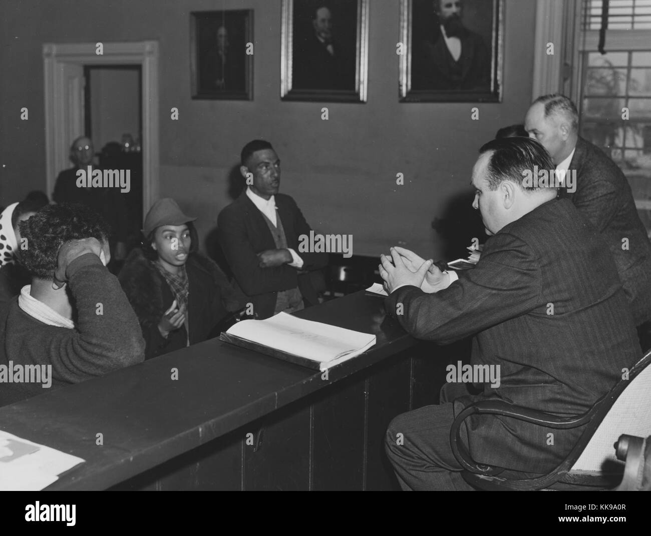 A photograph of a group of African American citizens appearing before two white court officials, everyone is dressed in nice clothing, the two groups are separated by a high wooden barrier and the officials are on an elevated platform, another man watches from a doorway, the room is decorated with three portraits of different men, Rustburg, Virginia, March, 1941. From the New York Public Library. Stock Photo