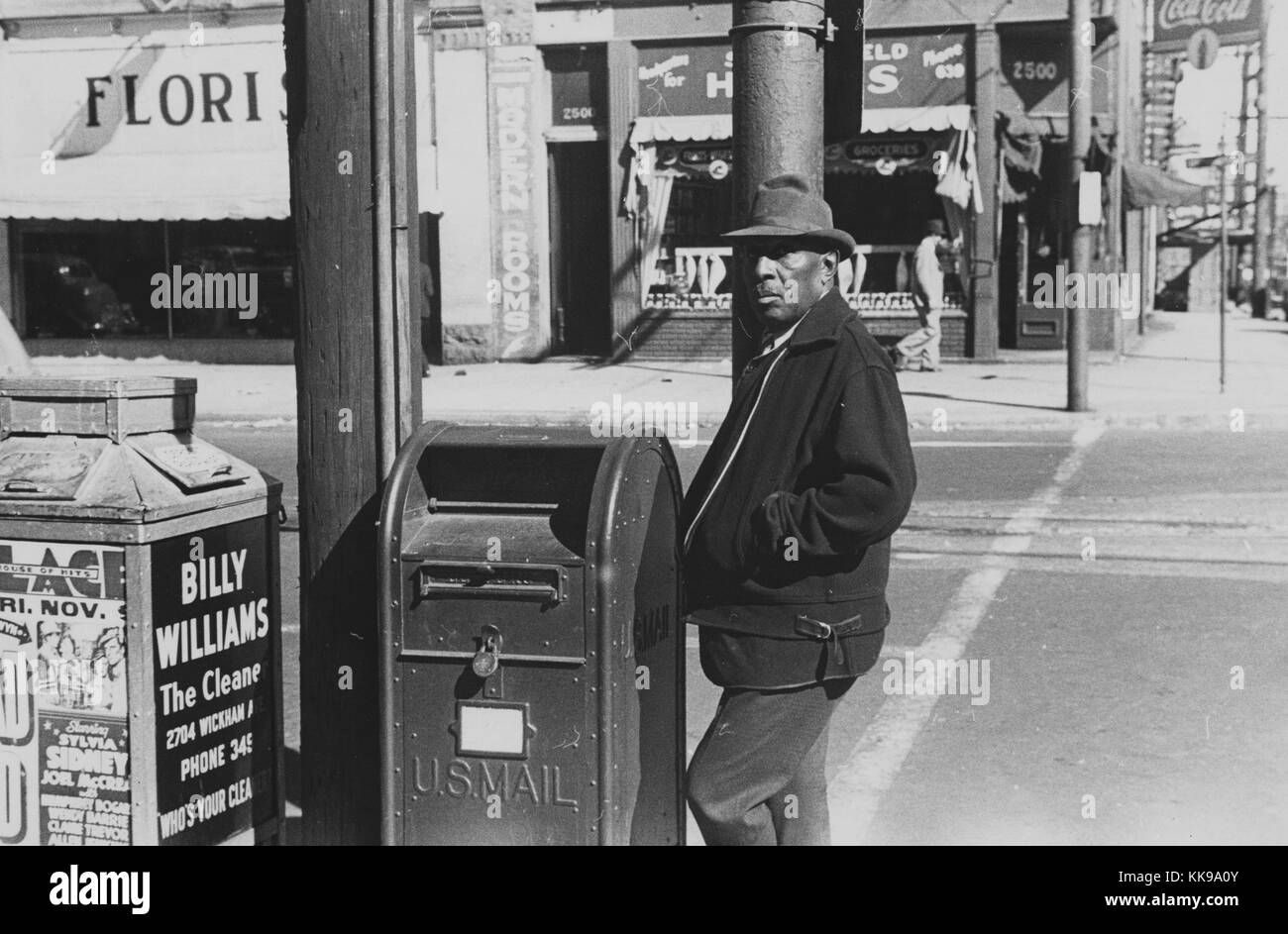 A photograph of a man standing by a United States Postal Service drop box on street corner, the man is wearing a dark colored jacket, pants and a hat, a trash bin is located next to the drop box and contains advertisements for local businesses and events, stores can be seen across the street directly behind the man, Newport News, Virginia, 1940. From the New York Public Library. Stock Photo