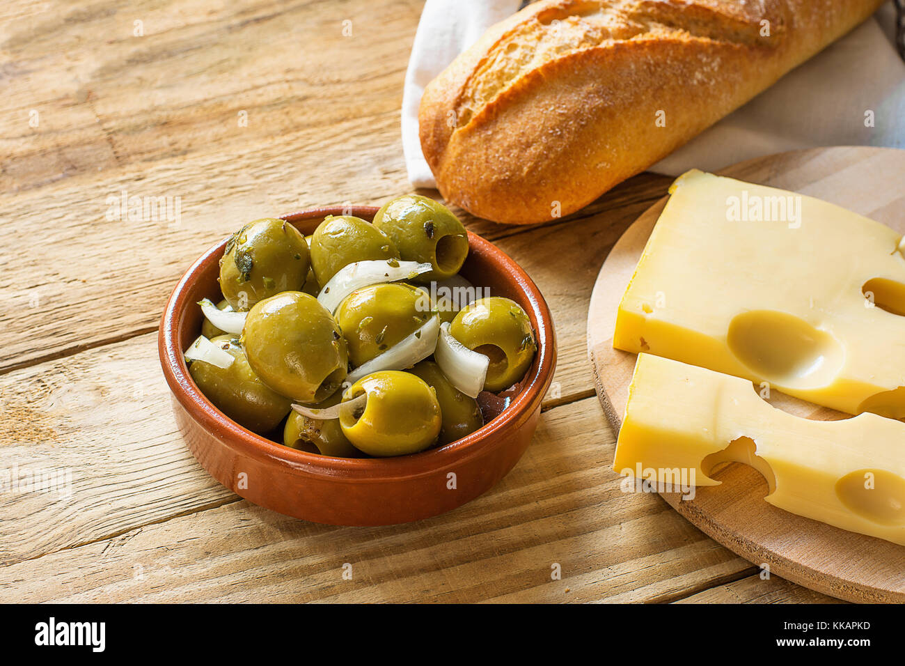 Big Spanish Green Gordal Olives with Herbs and Onions in Earthenware Bowl Baguette Maasdam Cheese on Cutting Board Wood Table Rustic Interior Mediterr Stock Photo