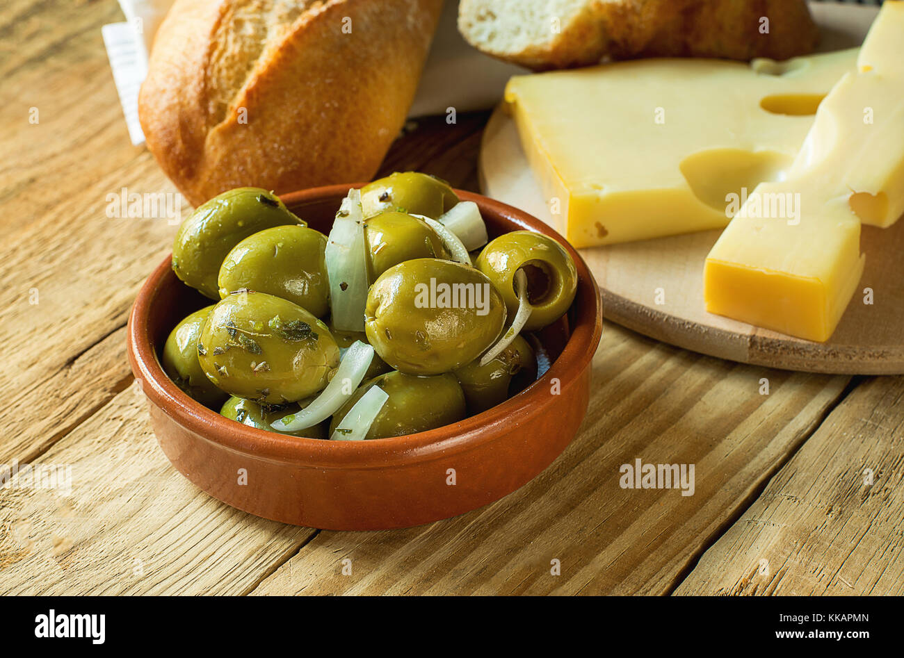 Big Spanish Green Gordal Olives with Herbs and Onions in Earthenware Bowl Sliced Baguette Maasdam Cheese on Cutting Board Wood Table Rustic Interior M Stock Photo