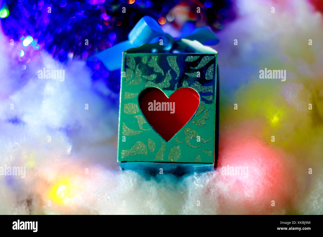 Close up blue gift box with red heart and blue ribbon on soft snowy sparkling background. Stock Photo