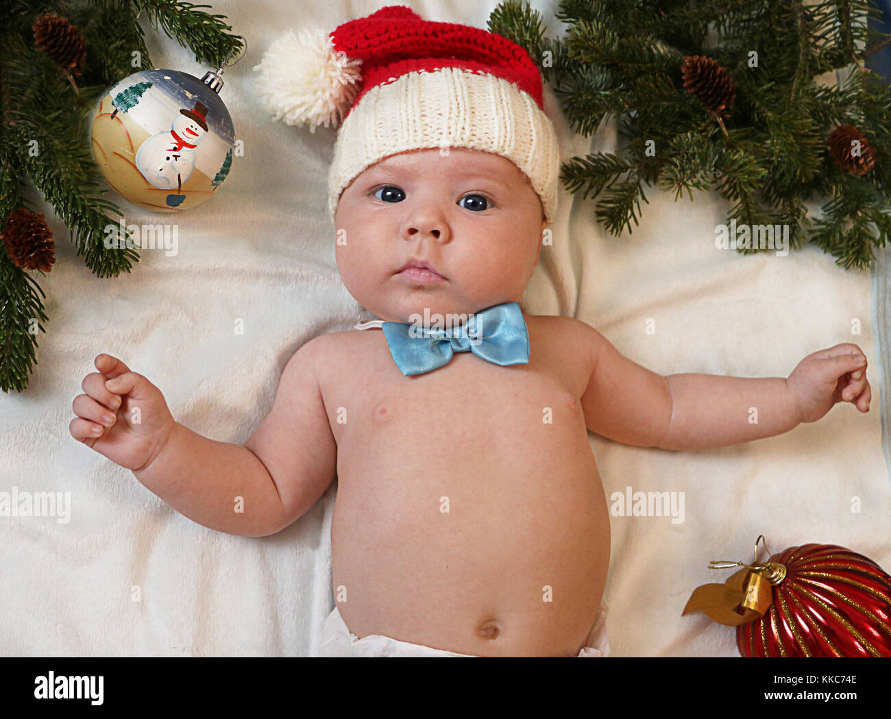 Baby first christmas. Beautiful little baby in Santa hat and bow tie lays on white background framed with christmas tree needles and balls. New Year's Stock Photo