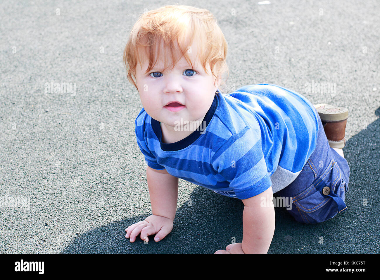 Fashioned ginger toddler boy with blue eyes staying on all fours on the play ground and watching on camera. Stock Photo
