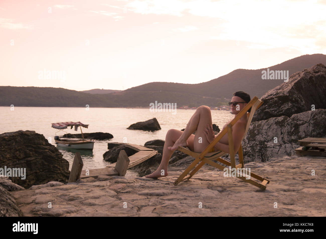 one young man relaxing sitting in beach chair, sea, old fishing boat, rocks. Jaz beach, Montenegro. Stock Photo