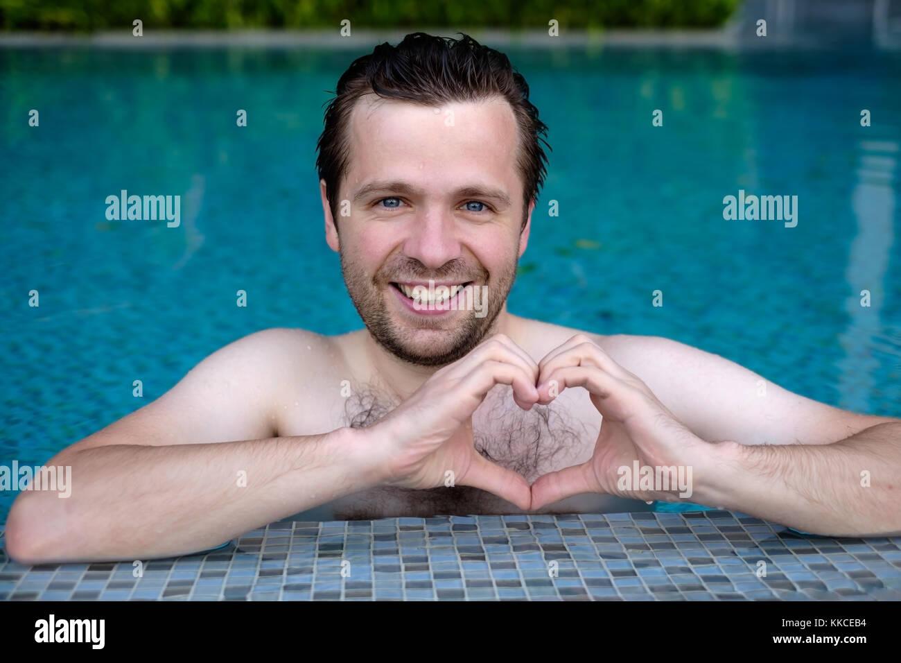 Handsome smiling young caucasian man makes the heart using fingers, hands during swimming at pool. Stock Photo