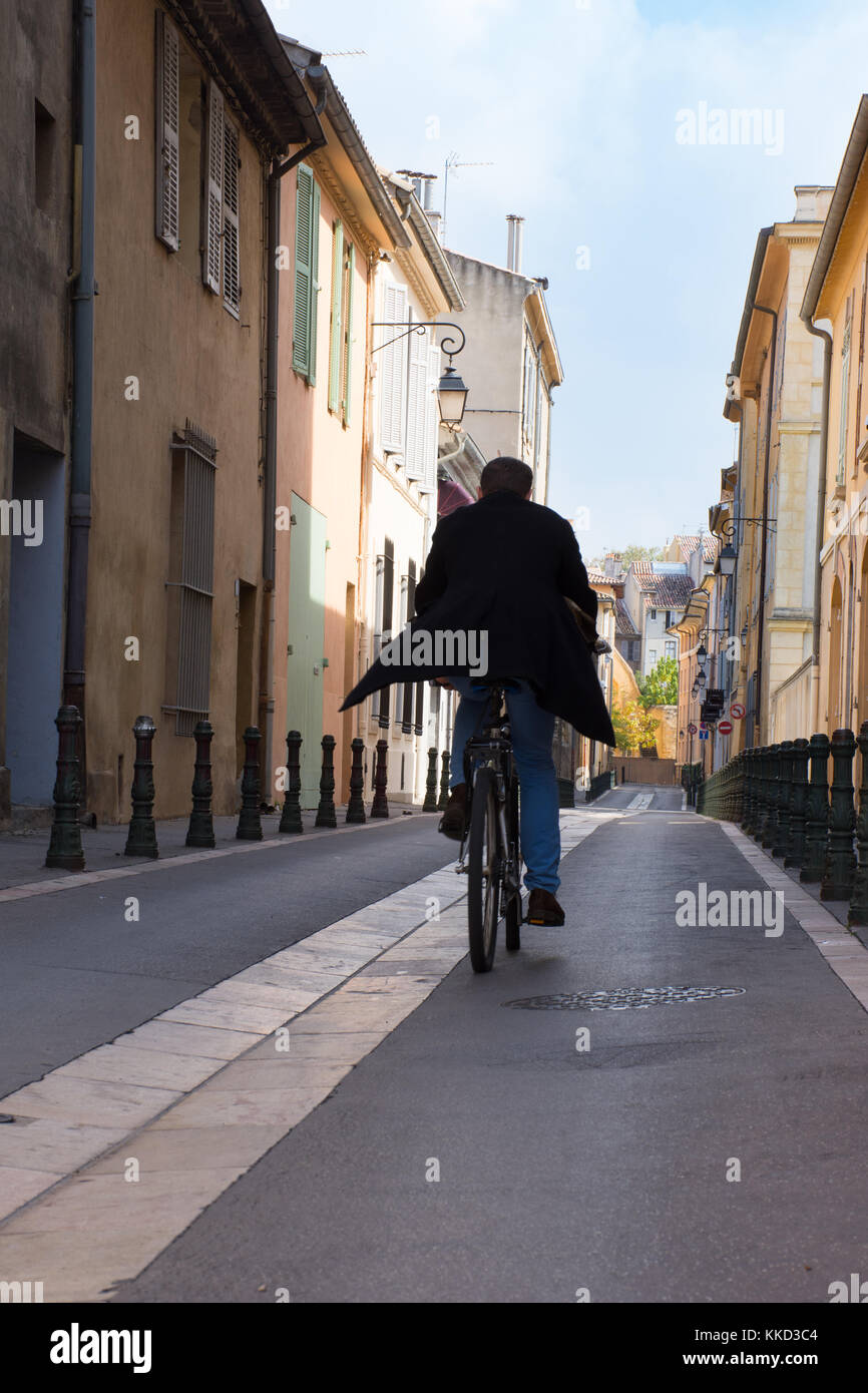 Bicyclist in jeans and a black coat on a narrow street lined with pastel stucco buildings. His back is to the camera. Stock Photo