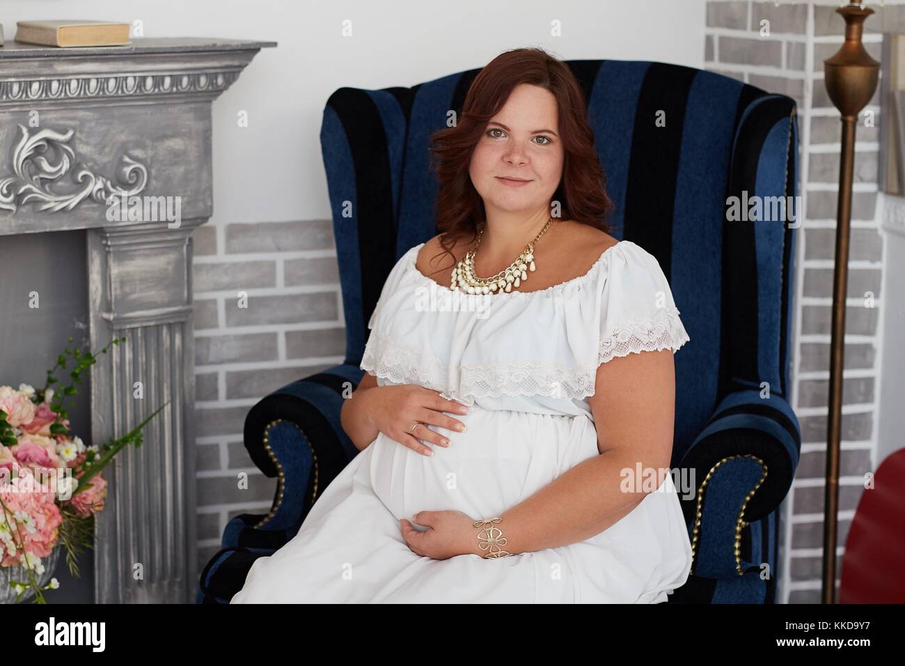 Portrait of adorable pregnant woman with happy smile in pure white dress and jewelry embraces her belly sitting in vintage armchair near to fireplace Stock Photo