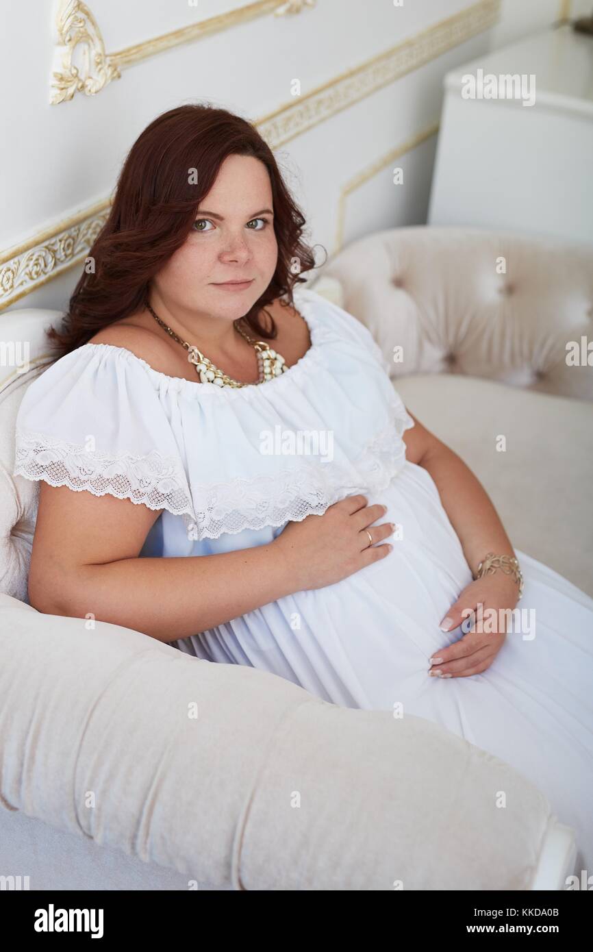Peaceful beautiful pregnant woman with happy smile in pure white dress and jewelry put her hand on belly sitting on white sofa in luxury decorated int Stock Photo