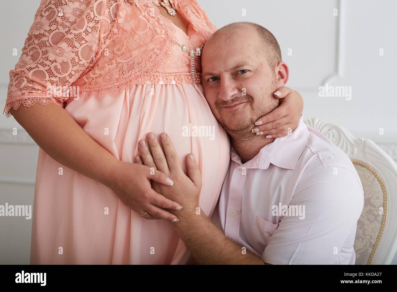 Man embracing belly of pregnant woman in coral dress  on luxury decorated white wall background. Stock Photo