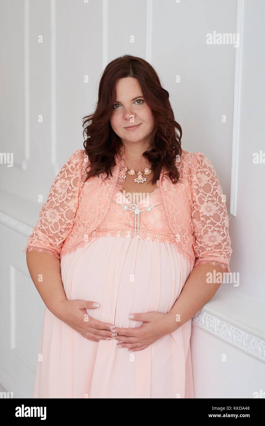 Beautiful pregnant woman with calm smiling  peaceful face dressed in coral dress and jewelry embracing her belly standing on white luxury decorated wa Stock Photo