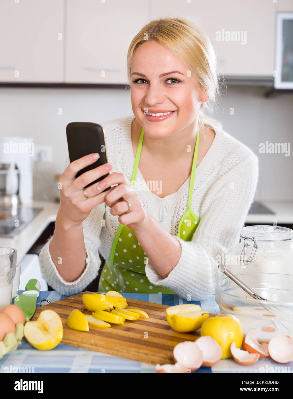 Young Smiling Blonde Housewife In Apron Reading Recipe At Smartphone