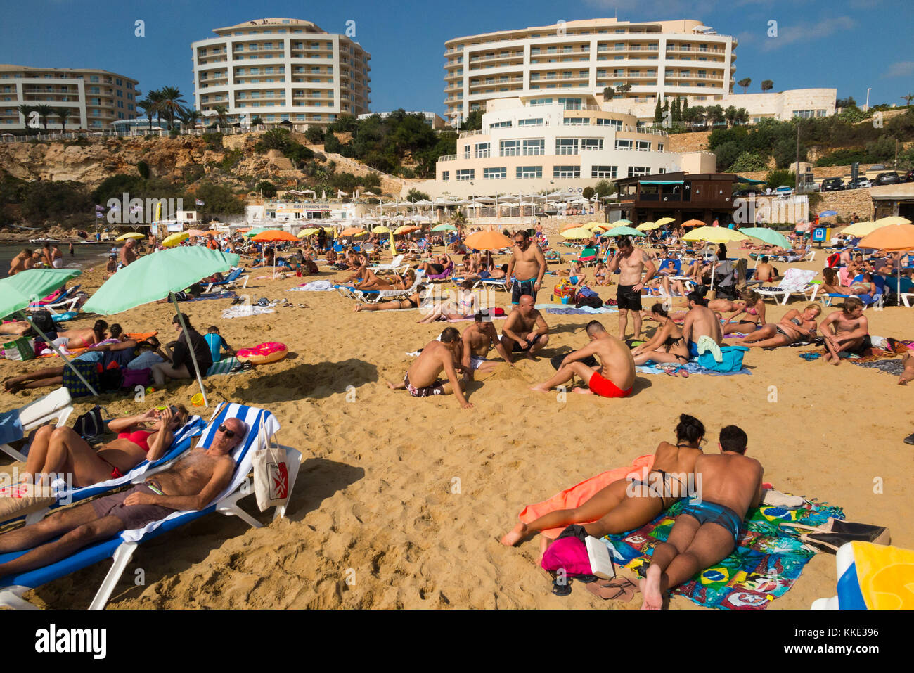 Sea & sandy beach with tourists / sunbathers people enjoying themselves swimming sun bathing overlooked by Radisson Blu Resort, Malta Golden Sands. Stock Photo
