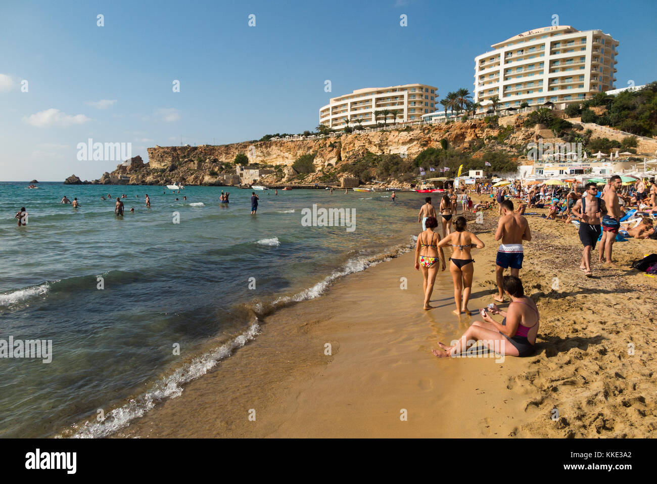 Sea & sandy beach with tourists / sunbathers people enjoying themselves swimming sun bathing overlooked by Radisson Blu Resort, Malta Golden Sands. Stock Photo