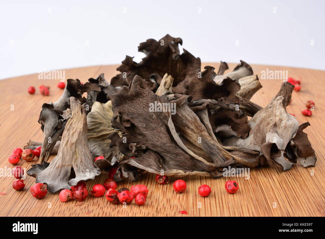 Pile of dried Horn of plenty or Craterellus cornucopioides mushrooms with spicy red pepper on bamboo wooden chopping board over white background Stock Photo