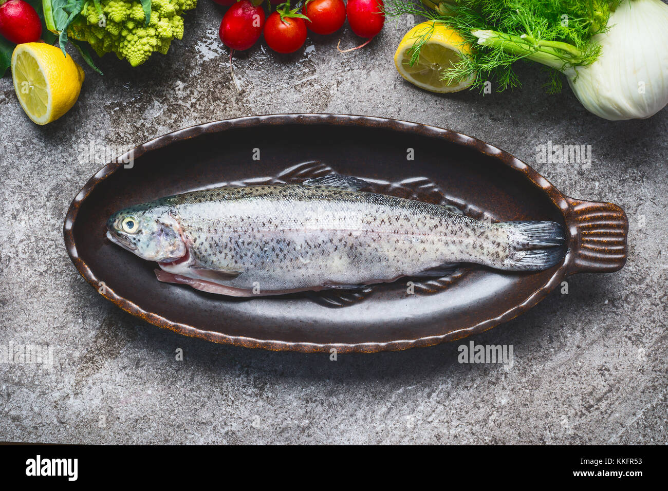 Raw fresh whole trout in baking dish with vegetables cooking  ingredients on gray concrete background , top view.  Healthy Food concept Stock Photo