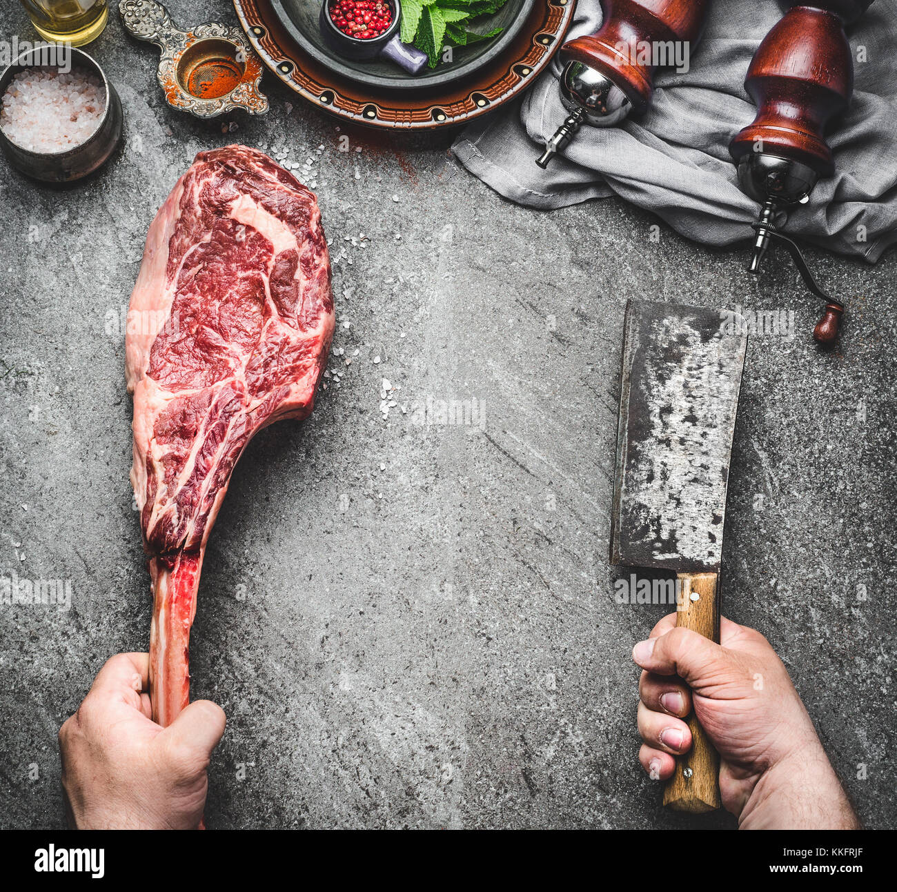 Male hands of  butcher or cook holding tomahawk beef steak and meat cleaver on dark rustic kitchen table background with cooking ingredients and condi Stock Photo