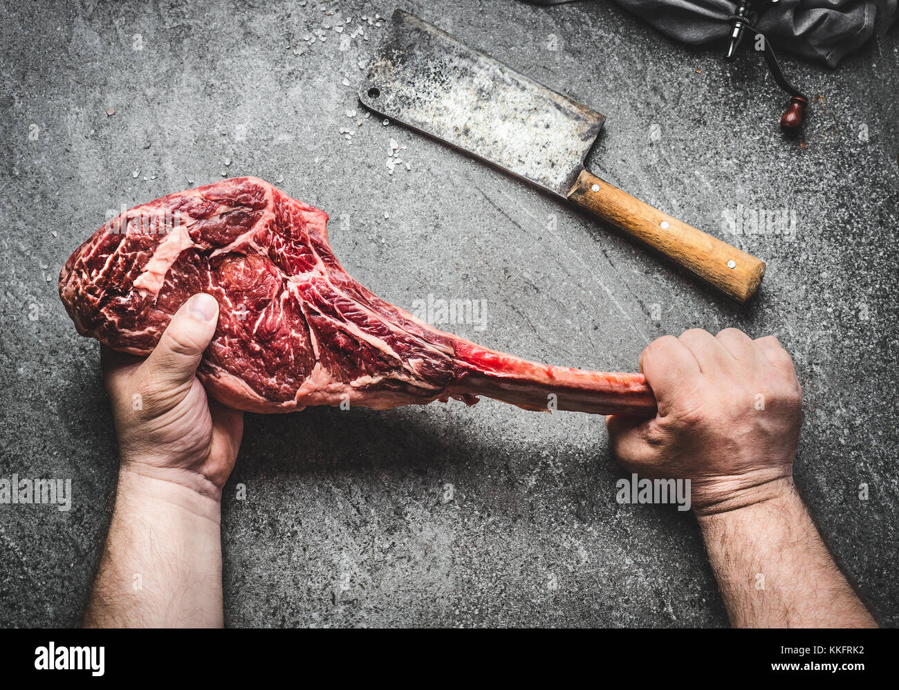 Raw tomahawk beef steak in male hands with meat cleaver on dark rustic background. Butcher hands holding tomahawk steak, top view Stock Photo