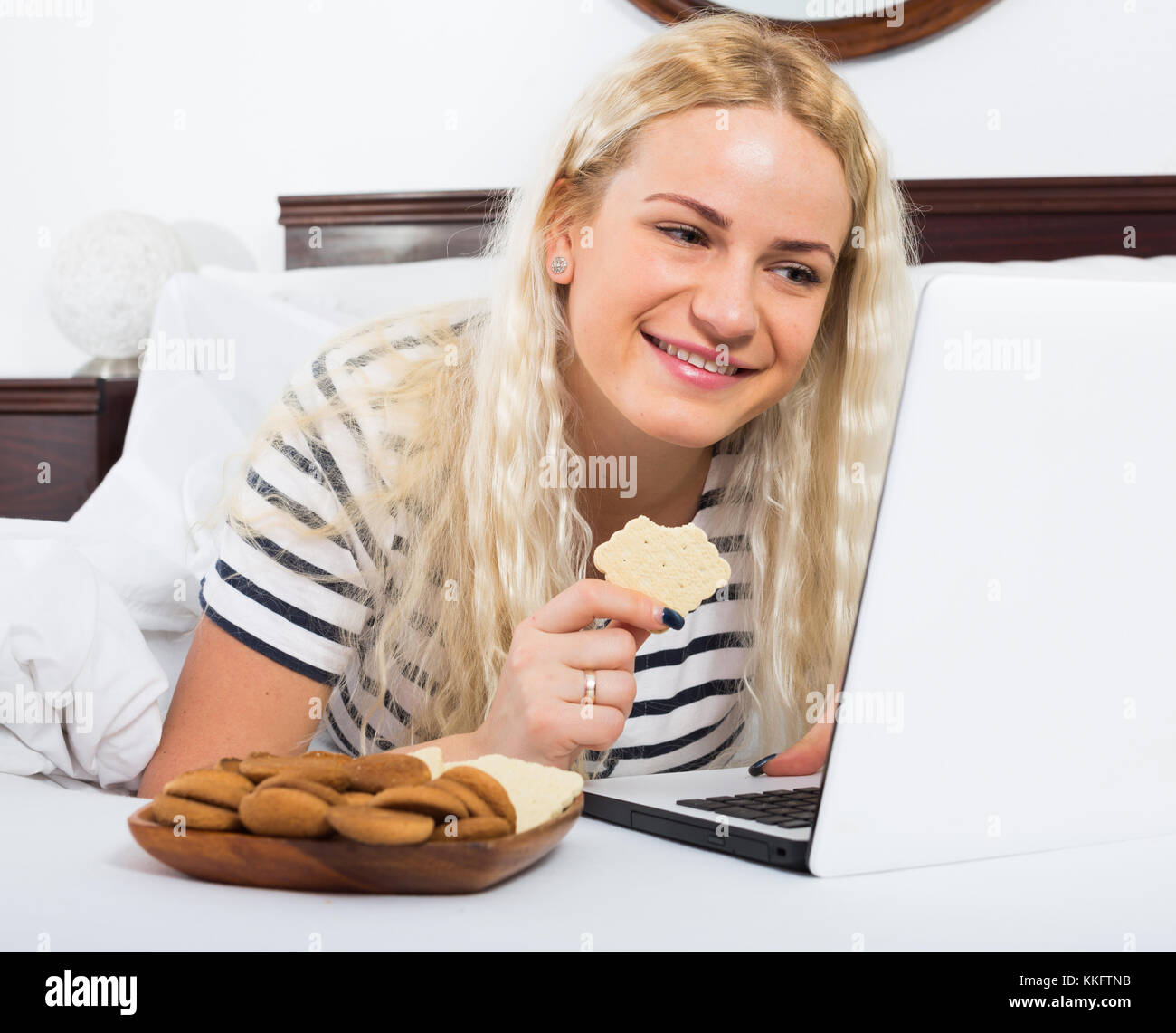Cheerful young woman with biscuits working on laptop in bed Stock Photo Alamy