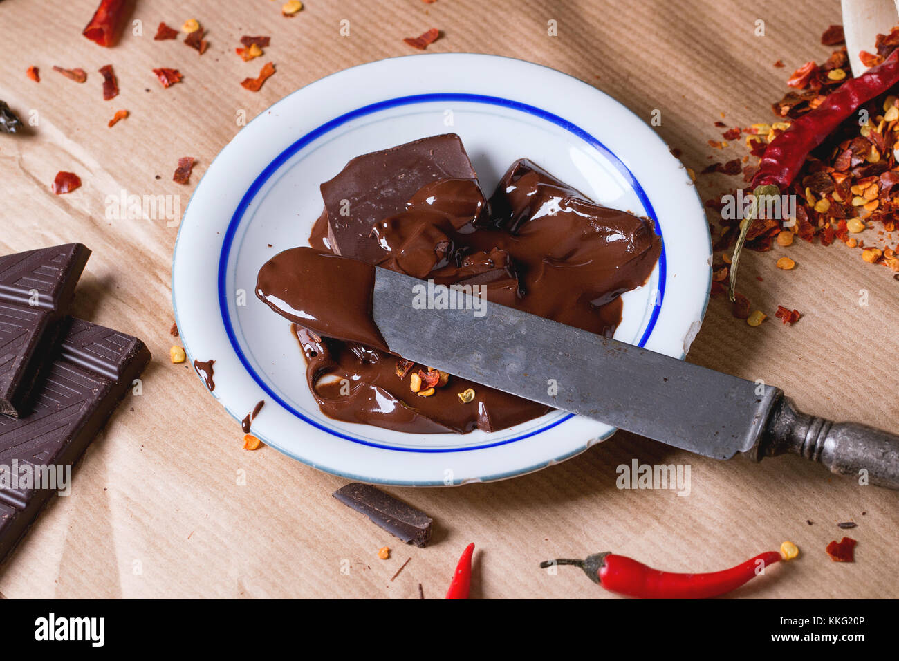 Chopping dark chocolate with fresh and dry red hot chili peppers on baking paper with plate of hot melted chocolate and vintage knife. Square image Stock Photo
