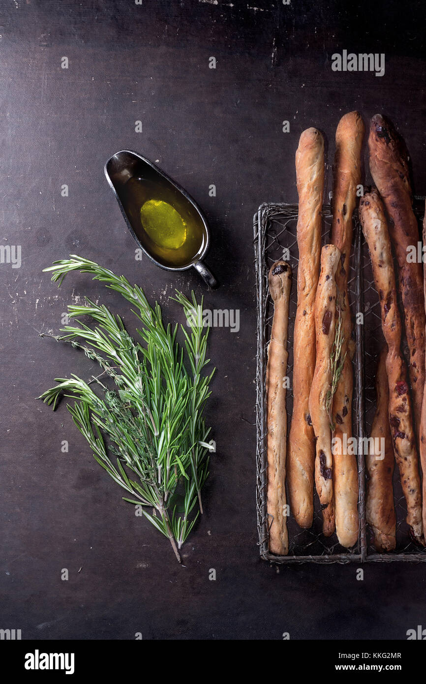 Fresh baked homemade grissini bread sticks in vintage metal grid box with olive oil and herbs rosemary and thym over dark surface. Top view. Stock Photo