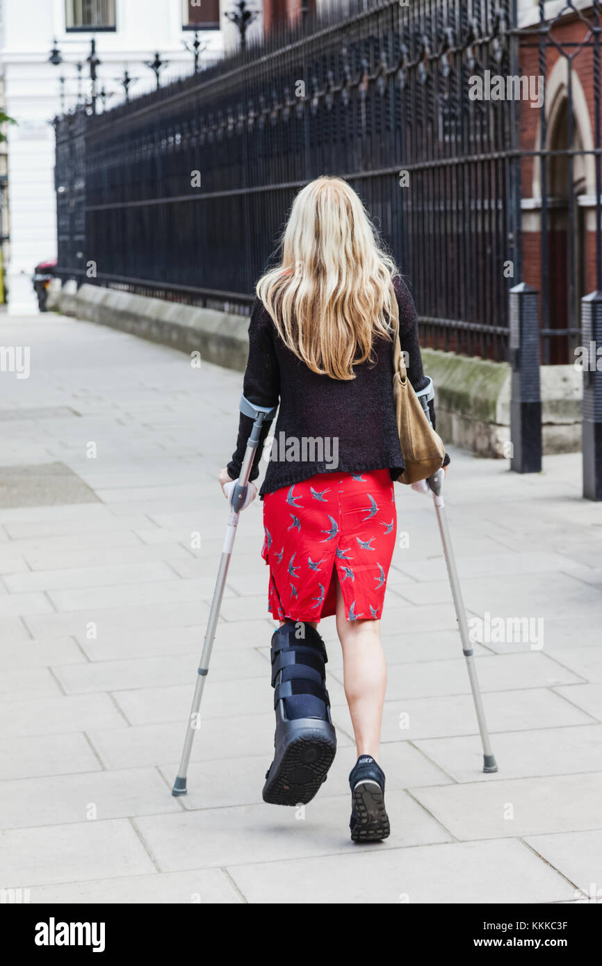 England, London, Young Lady Walking on Crutches in Street Stock Photo