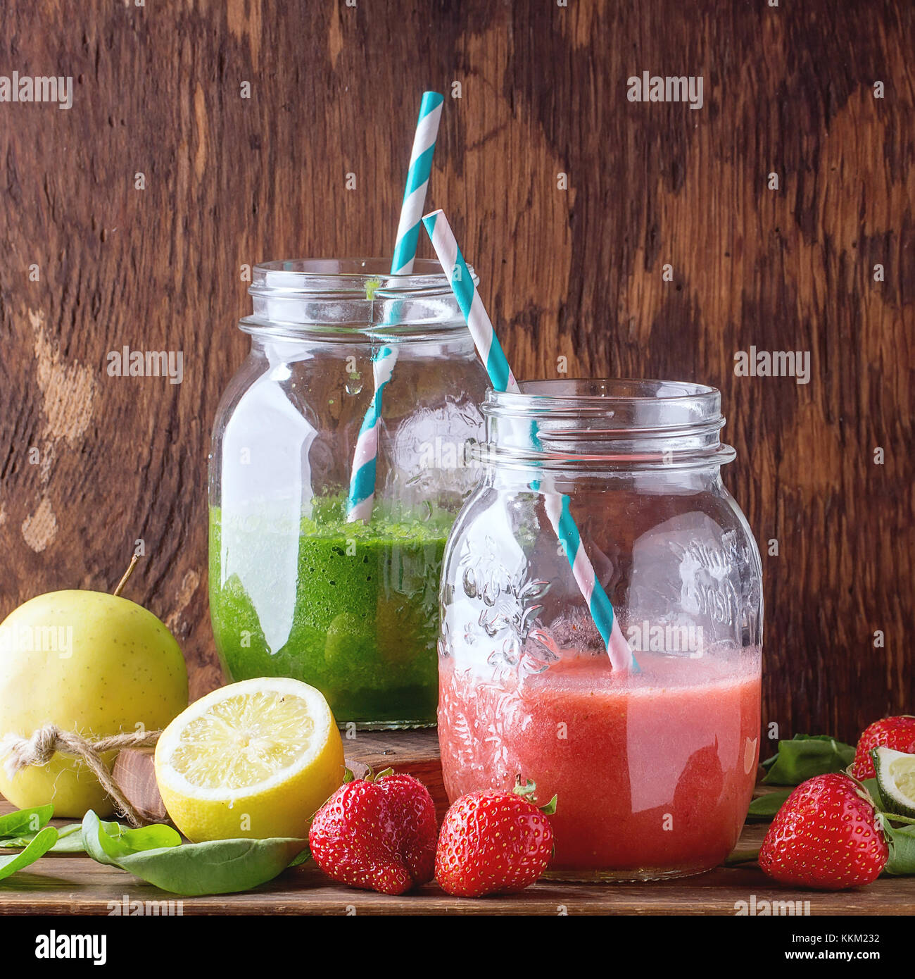 Glass jars with green and red smoothie, served with baby spinach, lemon, strawberries and green apple over wooden table. Square image with selective f Stock Photo
